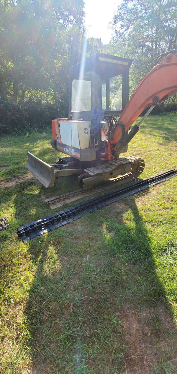A Kubota KH90 excavator with a missing rubber track sits on grass under sunlight; its detached track and Steel tracks Kubota KH90 are visible, along with trees in the background. | VIQAN Replacement Parts for Heavy Equipment - Rubber and Steel Tracks