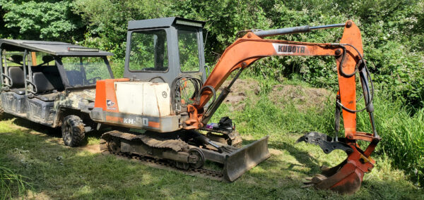 A Steel tracks Kubota KH90 is parked on grass beside a black utility vehicle, with green trees and bushes in the background. | VIQAN Replacement Parts for Heavy Equipment - Rubber and Steel Tracks