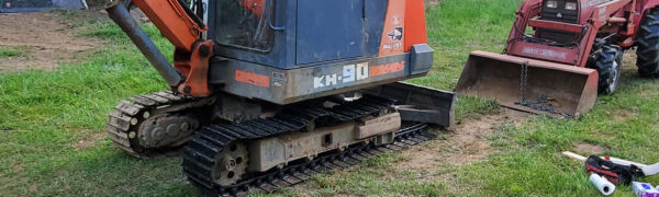 A Steel tracks Kubota KH90 excavator, compact and colored orange and blue, is parked on grass near a red tractor and scattered tools. | VIQAN Replacement Parts for Heavy Equipment - Rubber and Steel Tracks