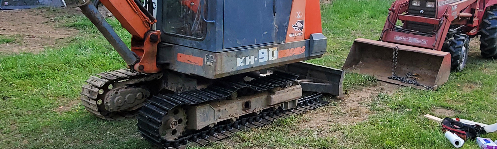 A Steel tracks Kubota KH90 excavator, compact and colored orange and blue, is parked on grass near a red tractor and scattered tools. | VIQAN Replacement Parts for Heavy Equipment - Rubber and Steel Tracks